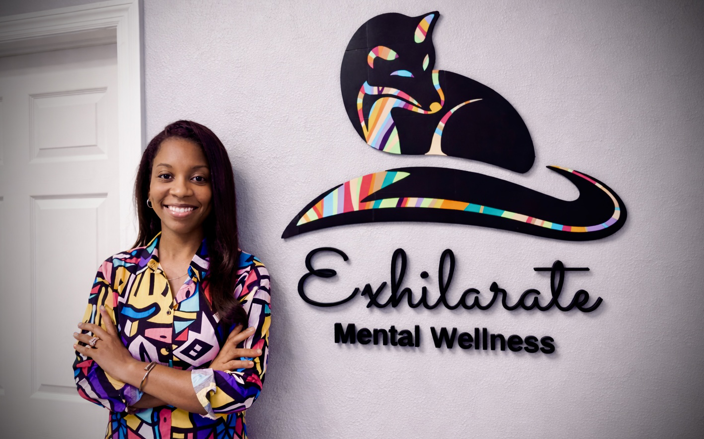 Amanda Baird in front of Exhilarate Mental Wellness sign in Tampa Bay, Florida offering play therapy services.